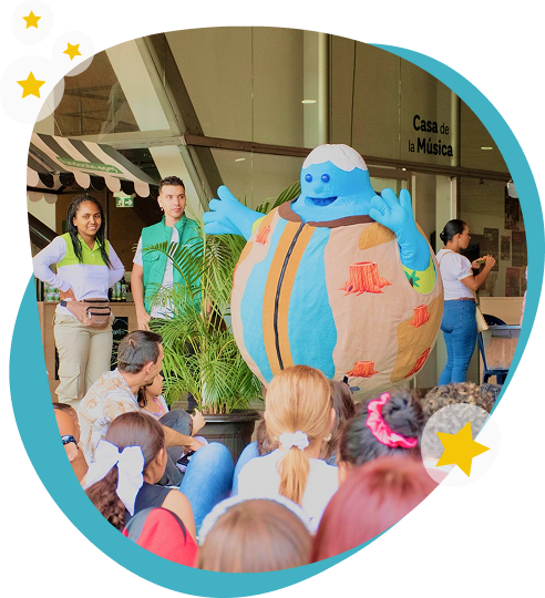 Actividad infantil con personaje animado en la Casa de la Música Niños sentados observan a un personaje animado azul en una actividad infantil frente a la Casa de la Música, acompañado por dos guías.