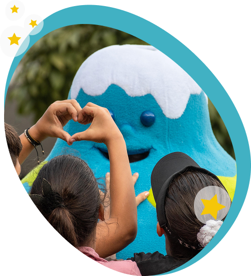 preguntas-frecuentes Niños sentados observan a un personaje animado azul en una actividad infantil frente a la Casa de la Música, acompañado por dos guías.